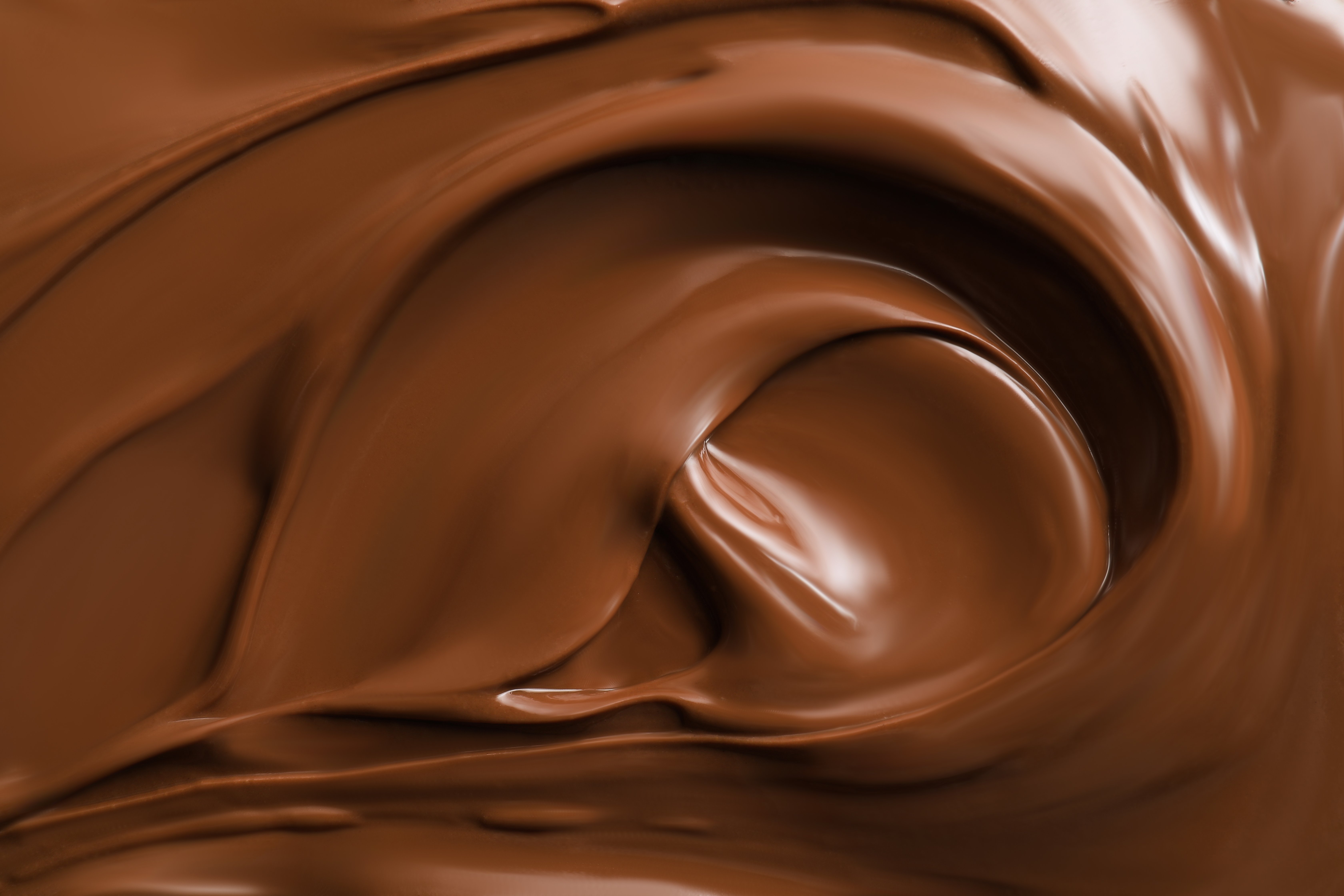 A confectioner pours smooth melted chocolate into a rectangular mold beside an industrial mixing machine. The scene takes place in a professional kitchen or candy shop with a vibrant purple wall in the background. Thick chocolate flows from the machine into a metal basin while the worker carefully levels the tray. The image emphasizes craftsmanship, rich textures, and the warm golden-brown color of the chocolate.